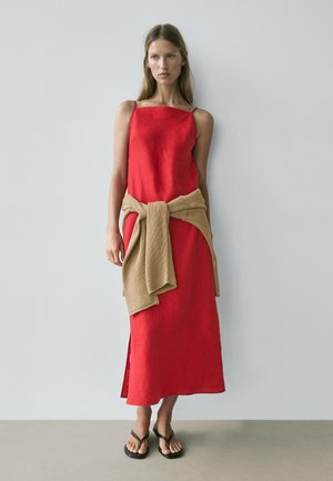 Giovane donna in abito midi rosso senza maniche con maglione beige annodato in vita e sandali infradito neri, in piedi contro una parete uniforme.