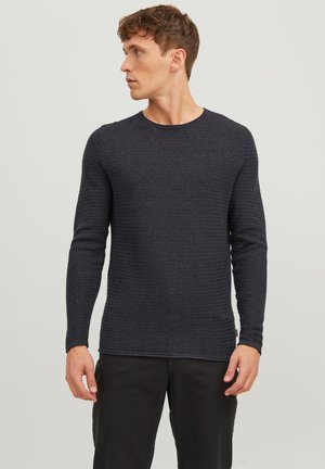 Hombre con un suéter de manga larga gris oscuro texturizado y pantalones negros, mirando hacia la izquierda contra un fondo claro y liso.