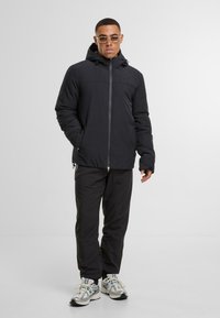 Sort isoleret jakke med lynlås foran, justerbar hætte og håndlommer, parret med sorte bukser og reflekterende sølv sneakers.