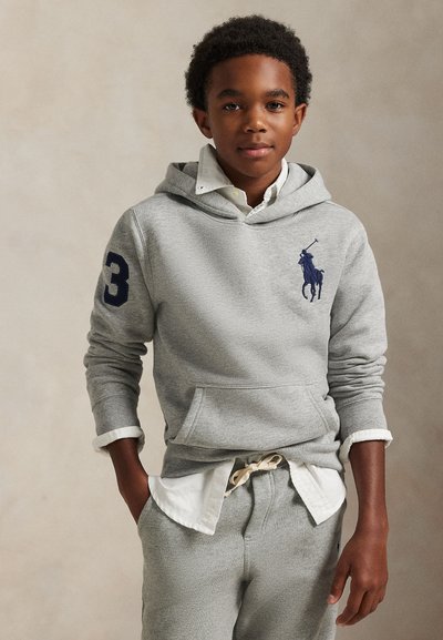 Garçon portant un sweat à capuche gris avec un logo de cheval bleu et le numéro trois sur la manche, chemise blanche en dessous et pantalon gris assorti.