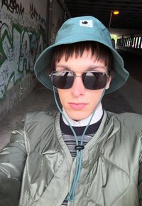Žalias kibiro tipo skrybėlė, pilkas siūlomas švarkas, juodi saulės akiniai, sluoksniuotas marškinys su raštuota tekstūra; miesto aplinka su grafičiais fone.