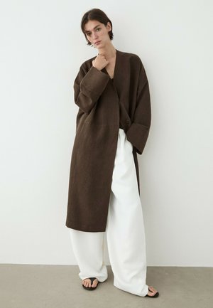 Vrouw draagt oversized donkerbruine jas, brede witte broek en zwarte sandalen, staand tegen een effen lichte muur met hand bij de kin.