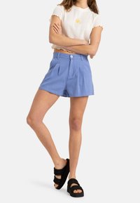 Pantaloni corti blu plissettati con chiusura a bottone, abbinati a un top corto bianco con texture e ricamo di un fiore giallo. Sandali neri con zeppa.