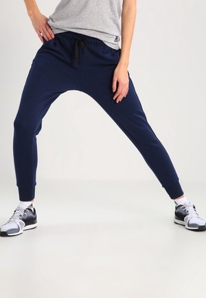 Osoba ubrana w granatowe spodnie dresowe i białe buty sportowe Adidas, stojąca z nogami szeroko rozstawionymi na jednolitym tle.
