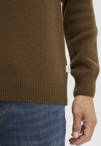 Pull en tricot marron avec des poignets et un ourlet côtelés. Étiquette visible sur le côté. Porté avec un jean en denim bleu, montrant un tissu texturé.