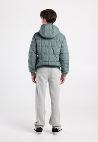 Groene donsjack met capuchon, voorzien van een doorgestikt design en geribbelde manchetten. Gecombineerd met grijze joggingbroek en zwarte sneakers.