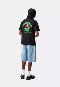 Camiseta de algodón negra con texto gráfico en negrita en verde y rojo en la parte posterior, combinada con pantalones cortos de mezclilla azul claro y zapatos negros.