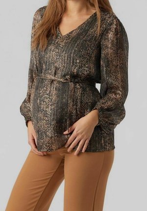 Gravid kvinde iført en brun mønstret bluse med taljebånd og beige bukser, som blidt holder sin mave med begge hænder.