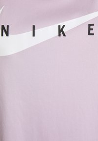 Camiseta deportiva de color lila con un logo curvado de Nike en blanco y letras negras en negrita. La tela tiene una textura suave.