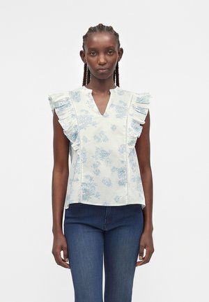 Mujer con camiseta blanca sin mangas con estampado floral azul claro y hombros con volantes, combinada con vaqueros azul oscuro, de pie sobre un fondo blanco.