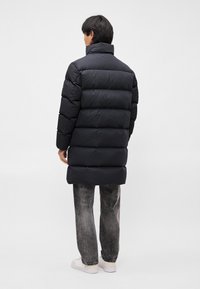 Schwarze Steppjacke mit hohem Kragen, ausgestattet mit horizontalen Steppnähten. Getragen mit grauen Jeans und weißen Sneakern, von hinten betrachtet.