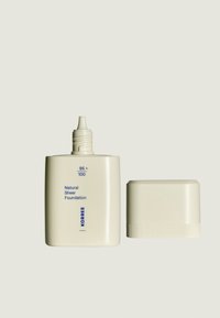 Natürliche, sheer Foundation in einer eleganten, matten Flasche in Off-White mit einer schmalen Ausgusstülle und einer passenden Kappe, die blaue Textdetails aufweist.