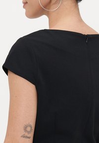 Svart kortermet kjole med rund hals og glidelås bak. Har en glatt tekstur og minimalistisk design.