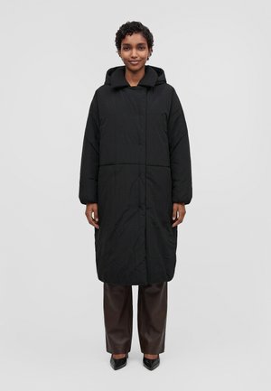Une femme porte un long manteau noir matelassé à capuche, un pantalon foncé et des chaussures noires sur un fond clair uni.