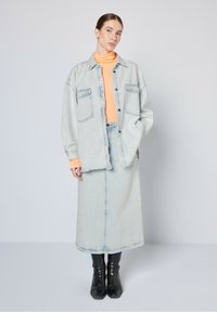 Giacca oversized in denim chiaro sopra un dolcevita arancione, abbinata a una gonna in denim coordinata. Stivali neri alla caviglia completano l'outfit.