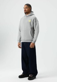 Grijze hoodie met een voorzak en een geel logo, gecombineerd met losse donkerblauwe jeans en zwarte schoenen met roze veters. Gladde stofstructuur.