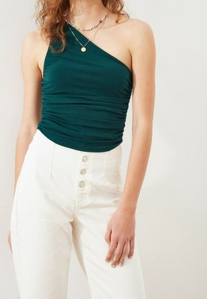 Kvinde iført en mørkegrøn off-shoulder top med drapering, hvide højtaljede bukser med knapper og lagdelte halskæder.