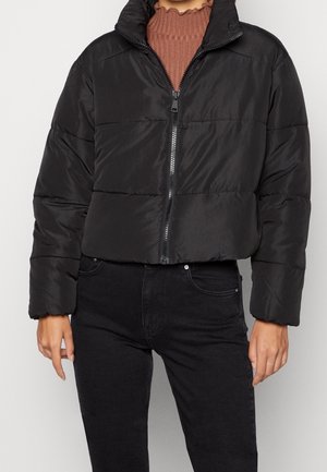 Femme portant une doudoune noire avec fermeture éclair, un haut côtelé marron et un jean noir, se tenant devant un fond uni.