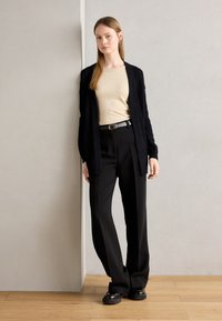 Černý pletený cardigan přes béžové tričko, v kombinaci s černými kalhotami s páskem. Černé boty, minimalistický design, hladká textura.