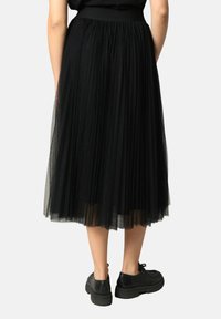 Jupe noire plissée en tulle, longue avec une taille élastique, associée à des chaussures noires à lacets, se tenant sur un fond neutre.