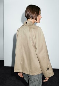 Veste beige surdimensionnée à la coupe structurée, subtilement brillante, avec des poignets à boutons, associée à un jean gris, vue de dos.