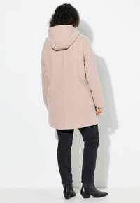 Abrigo capucha color rosa pálido, de corte recto, con mangas largas y bolsillos laterales, combinado con pantalones negros de corte slim y botines de tacón.