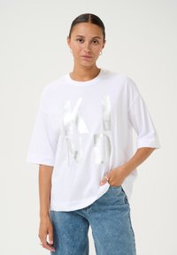 T-shirt oversize blanche avec un lettrage métallique argenté "KIND". Manches courtes, tissu en coton doux, coupe décontractée, idéale pour un look décontracté. Jean en denim.