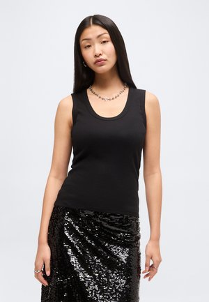 Młoda kobieta z długimi czarnymi włosami, ubrana w czarny bezrękawnik i błyszczącą czarną spódnicę z cekinami na jednolitym tle.