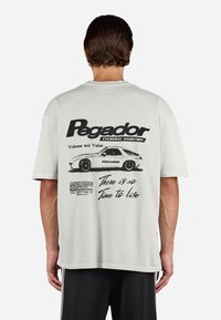 Man wearing white t-shirt with black vintage car graphic and text "Pegador," "Volume and Value," and "There is no time to lose" on the back.