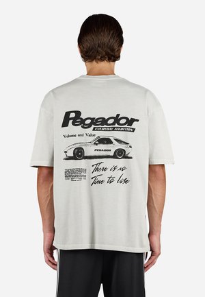 Man wearing white t-shirt with black vintage car graphic and text "Pegador," "Volume and Value," and "There is no time to lose" on the back.