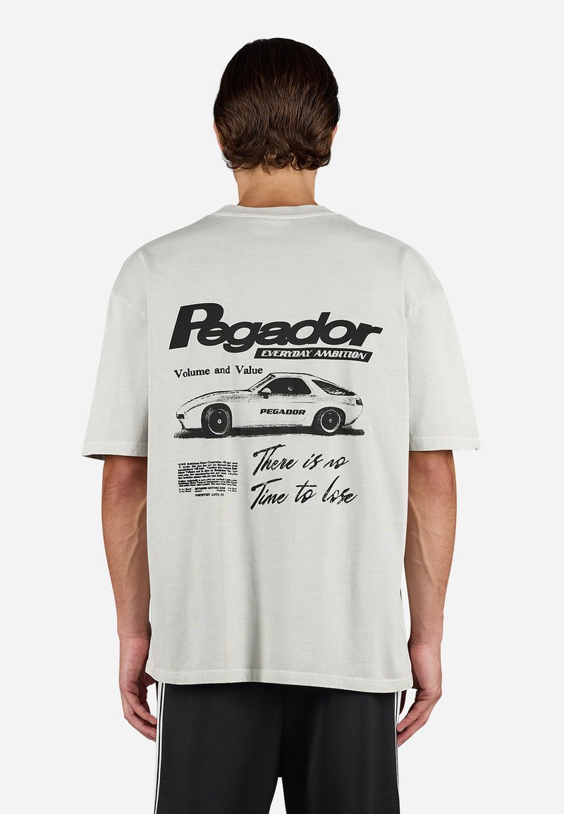 Man wearing white t-shirt with black vintage car graphic and text "Pegador," "Volume and Value," and "There is no time to lose" on the back.