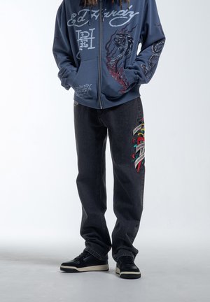 Person iført mørke jeans med farverig tatoveringslignende patch og marineblå lynlås-hættetrøje med drage- og tentakeldesigns.