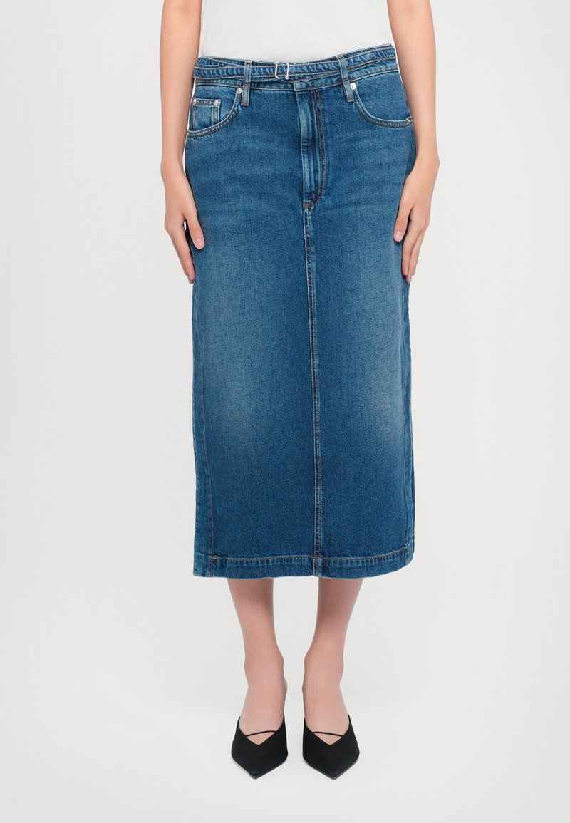 Jupe midi en denim bleu moyen, avec une coupe droite, des poches avant et un ourlet net. Associée à des chaussures noires à bout pointu.