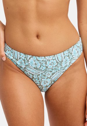 Femme portant un bas de bikini bleu et blanc à motif floral avec une taille basse, tenant la couture latérale d'une main.