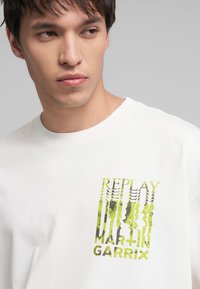 Biela bavlnená tričko s grafickým potlačou v zelenej a šedej farbe, zobrazujúca slová "REPLAY MARTIN GARRIX" so skresleným vzorom.