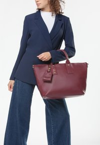 Sac fourre-tout en cuir bordeaux au design structuré, avec deux anses et une étiquette amovible. Le sac dispose d'accents en métal doré.