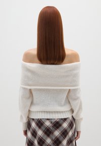 Suéter blanco, sin mangas y de corte off-the-shoulder, hecho de un material suave y peludo con puños acanalados. Combinado con una falda de cuadros que presenta tonos oscuros y claros.