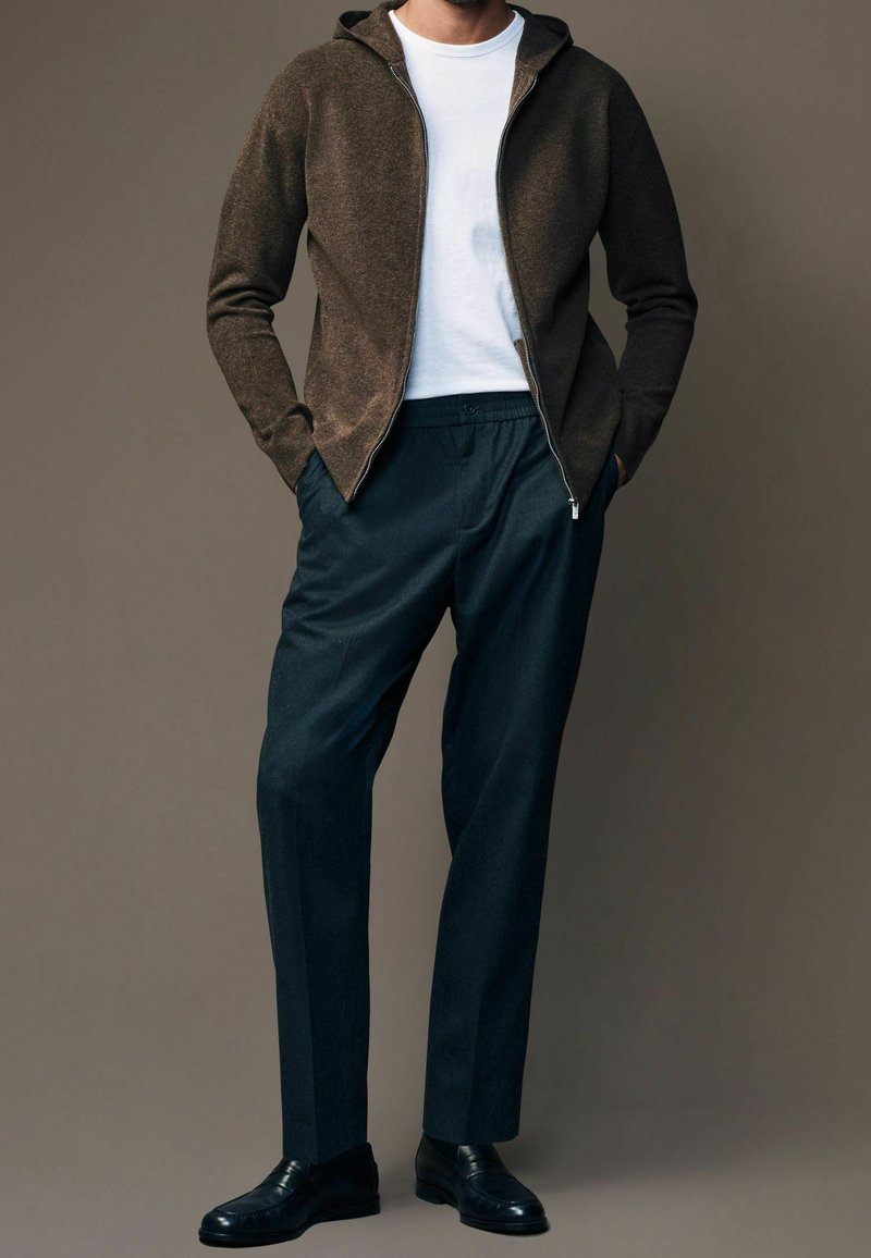 Homme portant un sweat à capuche zippé marron foncé sur un t-shirt blanc, pantalon bleu marine foncé et chaussures en cuir noires, debout les mains dans les poches.