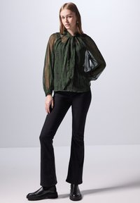 Blusa verde trasparente con collare alto, maniche lunghe e voluminose, e un motivo testurizzato. Abbinata a jeans neri a palazzo e stivali neri.