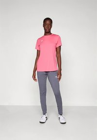 Rózsaszín sportpóló szürke melegítőnadrággal párosítva. A póló szabása normál; a melegítőnadrág szűkített. Mindkettőn az Under Armour logó található.