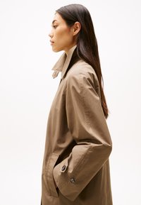 Cappotto trench in cotone marrone chiaro con un alto colletto, polsini abbottonati e tasche laterali, mostrato di profilo su uno sfondo neutro.