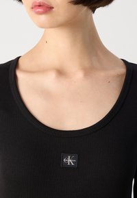 Zwarte geribbelde tanktop met een ronde hals en een Calvin Klein logo patch in het midden. Glad textuur, eenvoudig ontwerp, veelzijdige stijl.