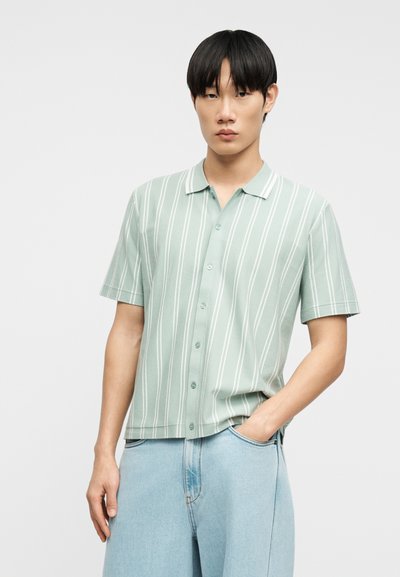 Νεαρός άνδρας που φοράει ανοιχτοπράσινο πουκάμισο με κοντά μανίκια και λευκές κάθετες ρίγες, μαζί με ανοιχτό μπλε τζιν, ποζάροντας με το ένα χέρι στην τσέπη.