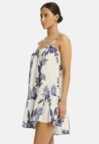 Das Modell in einem weißen Blumenkleid mit blauen Mustern steht seitlich und präsentiert den gerafften Saum des Kleides sowie die Spaghettiträgerr.