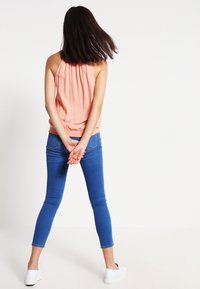 Top de tirantes color melocotón con tela texturizada, combinado con jeans ajustados azules y zapatillas blancas. La modelo está de espaldas, con las manos detrás de la espalda.