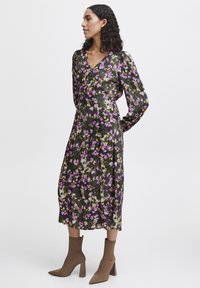 Robe florale vert foncé avec des accents violets et jaunes, dotée d'un décolleté en V, de manches longues et d'une longueur midi. Portée avec des bottes à talons marron.