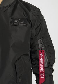 Svart bomberjacka i slät nylon med ett märke märkt "ALPHA INDUSTRIES," en dragkedjeficka och röd "REMOVE BEFORE FLIGHT"-tag.