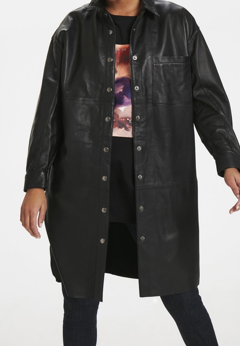 Personne portant un long manteau en cuir noir boutonné par-dessus un haut multicolore et un pantalon foncé, debout avec les bras légèrement étendus.