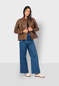Giacca cropped in pelle marrone con colletto a camicia, bottoni frontali e tasca, abbinata a pantaloni in denim blu a gamba larga e scarpe di colore chiaro.