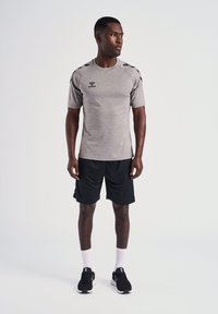 T-shirt de sport gris avec des accents noirs sur les épaules, associé à un short noir. Le modèle porte des baskets noires et des chaussettes blanches.